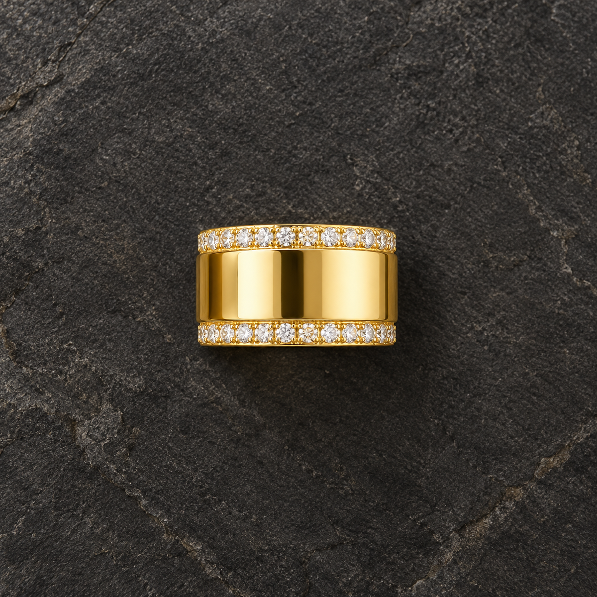 A wide yellow gold ring with pavé-set clear stones on both sides, centered on a dark textured stone surface.