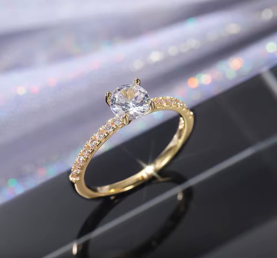A gold solitaire engagement ring with a round clear center stone and a pavé-set band of small clear stones, displayed on a glossy black surface with a soft iridescent blurred background.
