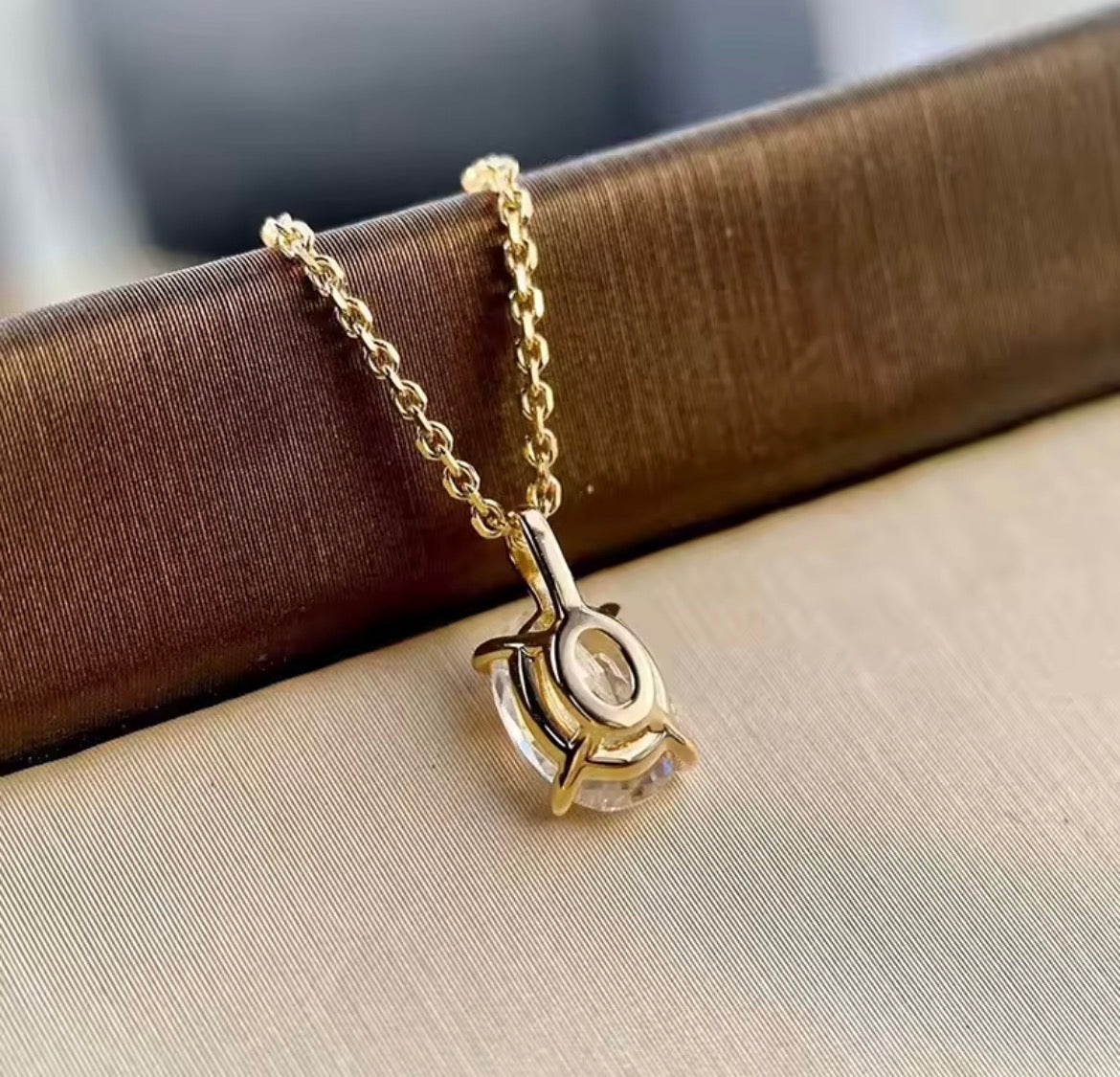 A gold chain necklace with a gold pendant base, displayed on a textured brown surface against a beige fabric background.