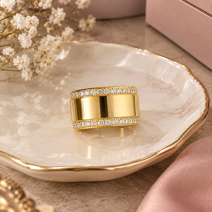 A wide gold ring with two rows of small clear stones, displayed on a white marble dish with gold trim. The scene includes soft pink fabric, white baby's breath flowers, and a pink box in the background.