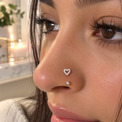 Close-up of a person's nose wearing two small silver nose studs, one heart-shaped stud and one small round stud, with a softly blurred indoor background.