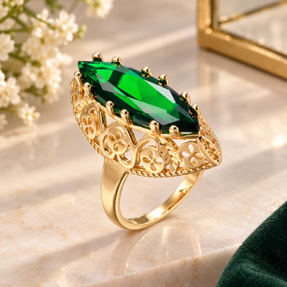 A gold ring with a large marquise-cut green gemstone, featuring ornate filigree detailing on the shoulders and a polished band, displayed on a light marble surface with soft-focus decorative flowers in the background.