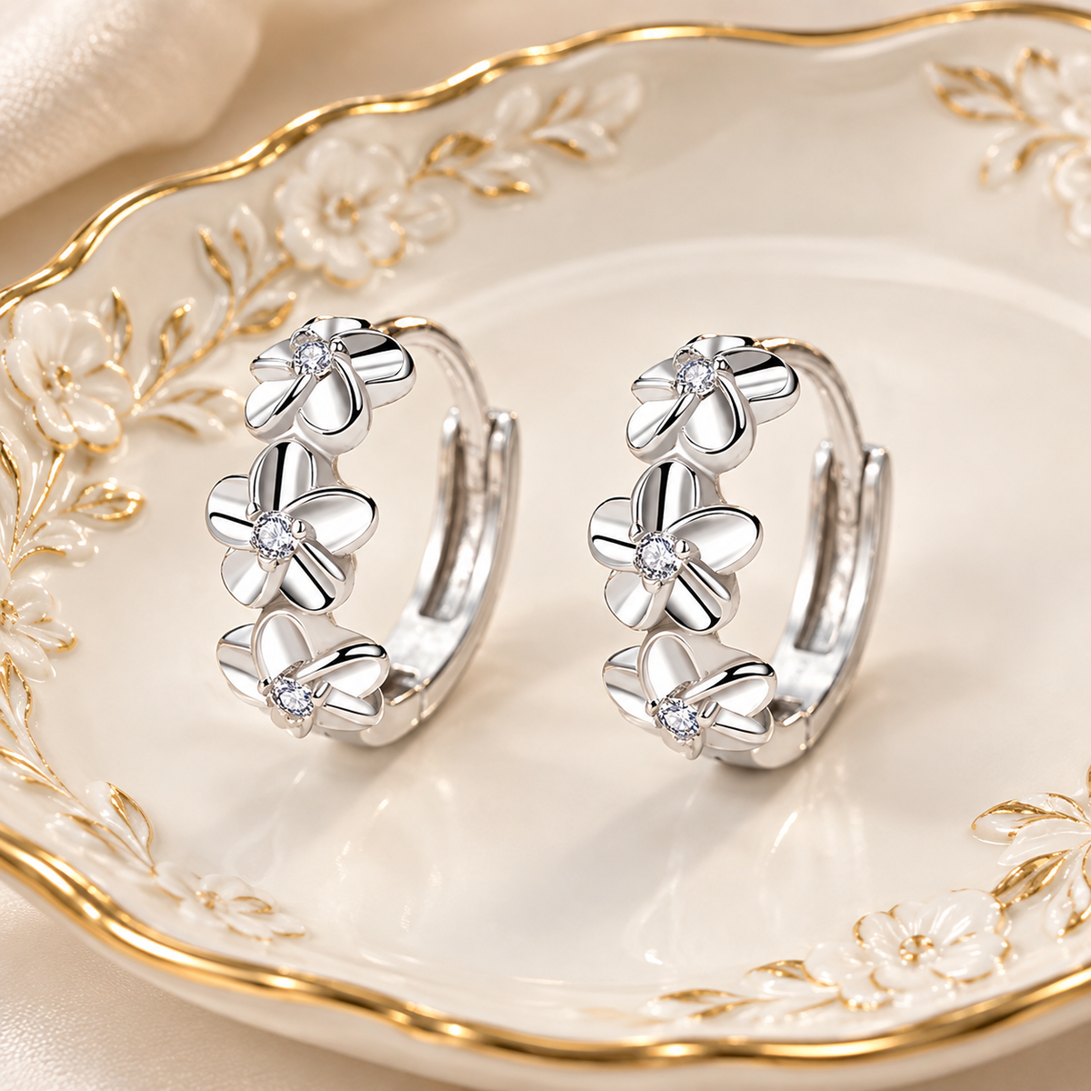 A pair of silver hoop earrings with three flower charms and small clear stones, displayed on a white ceramic dish with gold floral detailing.