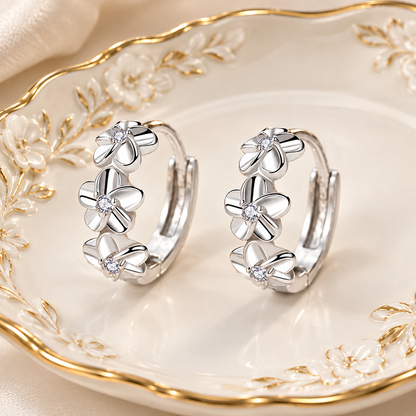 A pair of silver hoop earrings with three flower charms and small clear stones, displayed on a white ceramic dish with gold floral detailing.