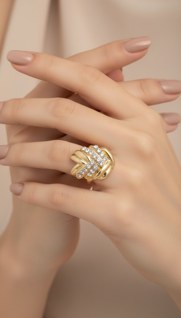 A close-up of a hand wearing a gold ring with a twisted knot design set with small clear stones, against a soft beige background.