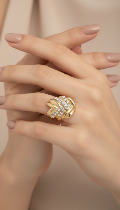 A close-up of a hand wearing a gold ring with a twisted knot design set with small clear stones, against a soft beige background.