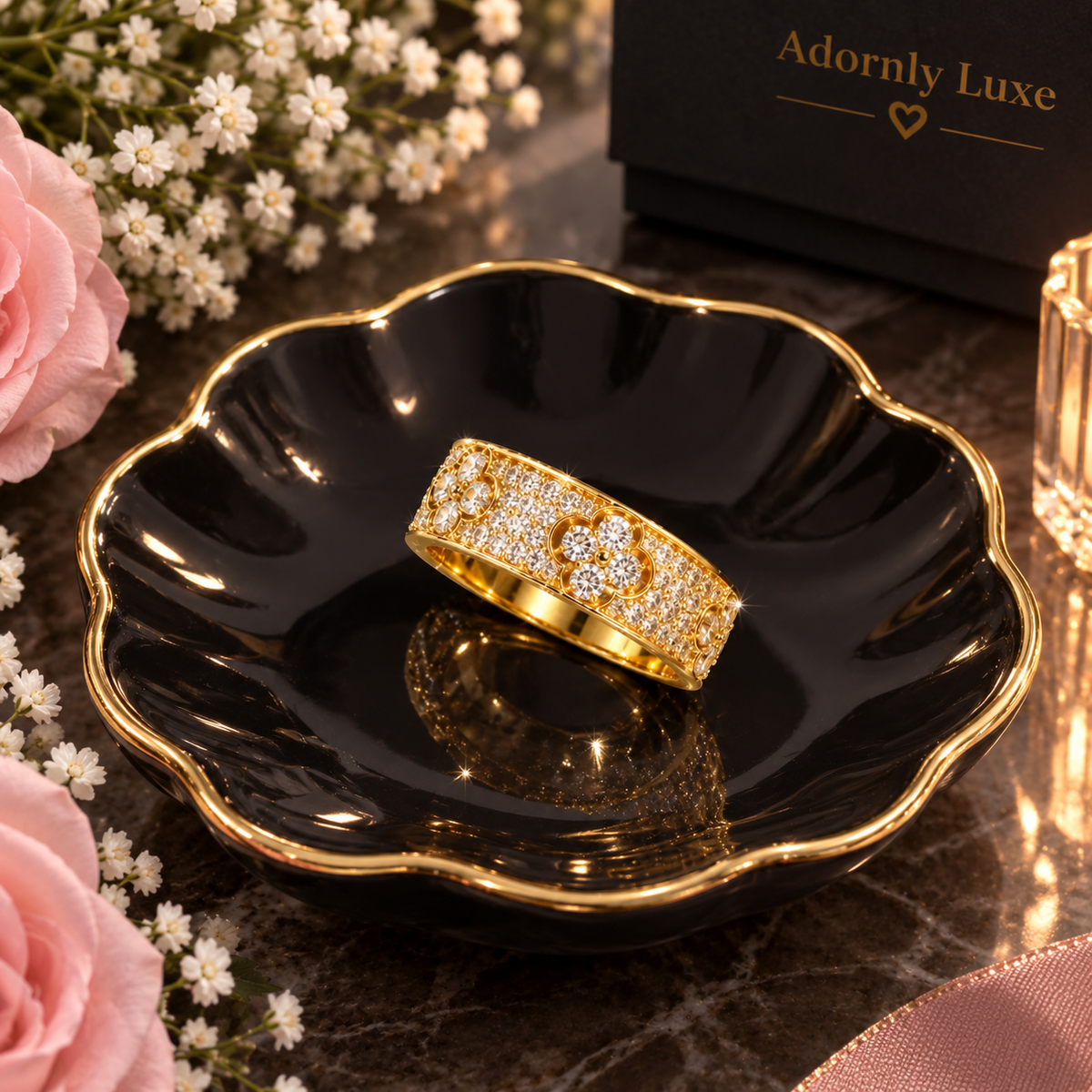 A gold ring with a floral pavé design sits on a black scalloped dish with gold trim. The scene is styled with pink roses, white baby's breath flowers, and a black gift box in the background that reads 'Adornly Luxe'.