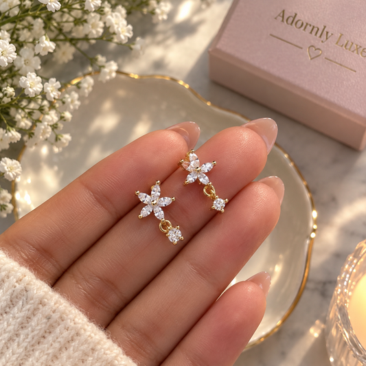 Two gold-tone flower jewellery pieces with clear stones and dangling accents, displayed on a hand beside a pink jewellery box.