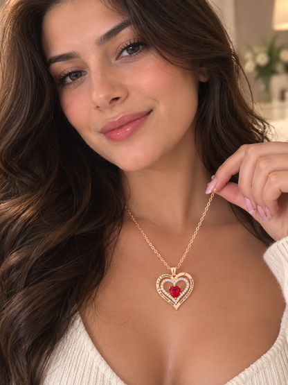 A woman wearing a gold heart pendant necklace with a red heart in the center and small clear stones around it, photographed in a softly lit indoor setting.