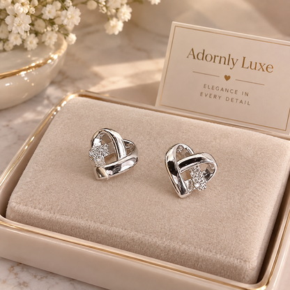 A pair of silver heart-shaped stud earrings with clear crystal details, presented in a beige jewelry box with a card that reads 'Adornly Luxe'.