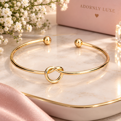 A polished gold cuff bracelet with a central knot design and small bead ends, displayed on a white marble tray with pink satin, baby's breath flowers, and a pink box labeled 'ADORNLY LUXE' in the background.