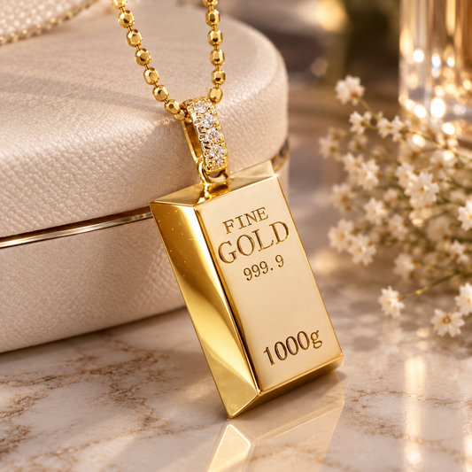 A gold bar pendant necklace engraved with 'FINE GOLD 999.9 1000g' hangs from a gold ball chain beside a cream-colored textured jewelry box on a marble surface with small white flowers in the background.