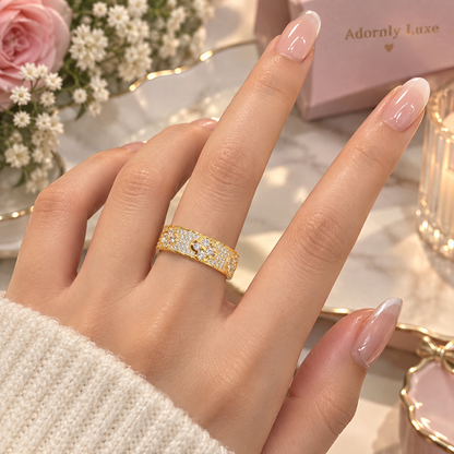 A close-up of a hand wearing a gold band ring set with small clear stones, styled with a pink gift box and flowers in the background. The box shows the brand name 'Adornly Luxe'.