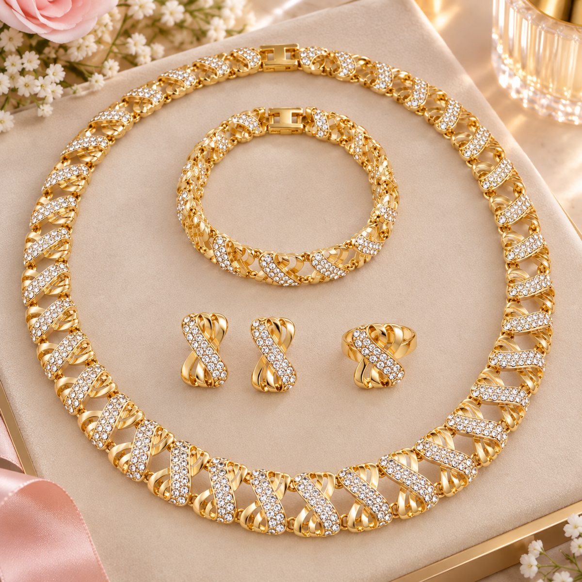 A gold-tone jewelry set displayed on a beige jewelry box, featuring a chunky Cuban link chain necklace, a matching bracelet, two matching earrings, and a ring, all adorned with clear rhinestones. The set is styled with pink roses and white flowers.