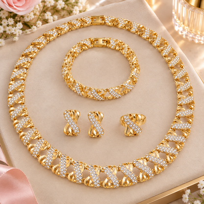 A gold-tone jewelry set displayed on a beige jewelry box, featuring a chunky Cuban link chain necklace, a matching bracelet, two matching earrings, and a ring, all adorned with clear rhinestones. The set is styled with pink roses and white flowers.