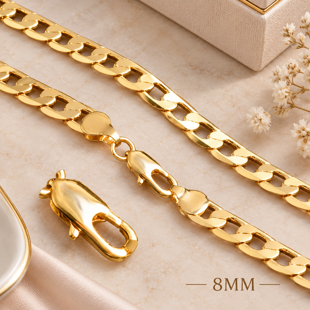 A close-up of a shiny gold chain necklace with a lobster clasp, laid on a light beige surface with decorative dried flowers and a small jewelry box. The text '8MM' appears near the bottom right.