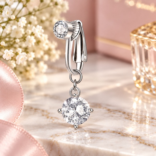 A silver-toned drop hoop earring with a large clear round gemstone dangle, photographed on a pink marble surface with soft floral and ribbon props.