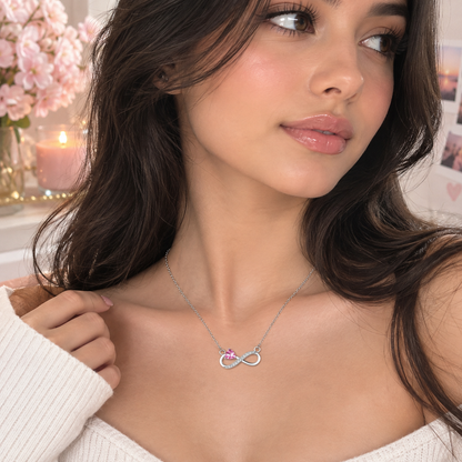 A woman wearing a delicate silver infinity necklace with a small pink gemstone accent, styled with a white ribbed knit top in a softly lit indoor setting.