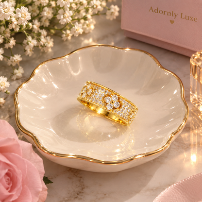 A gold ring with a floral pavé design sits in a white scalloped dish with gold trim. The scene is styled with a pink rose, white baby's breath flowers, a pink gift box labeled 'Adornly Luxe', and a glass tumbler on a marble surface.