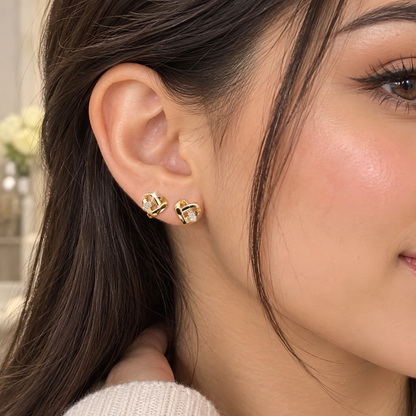 A close-up of a woman's ear wearing a pair of gold heart-shaped stud earrings with small clear stones, shown against her hair and skin.
