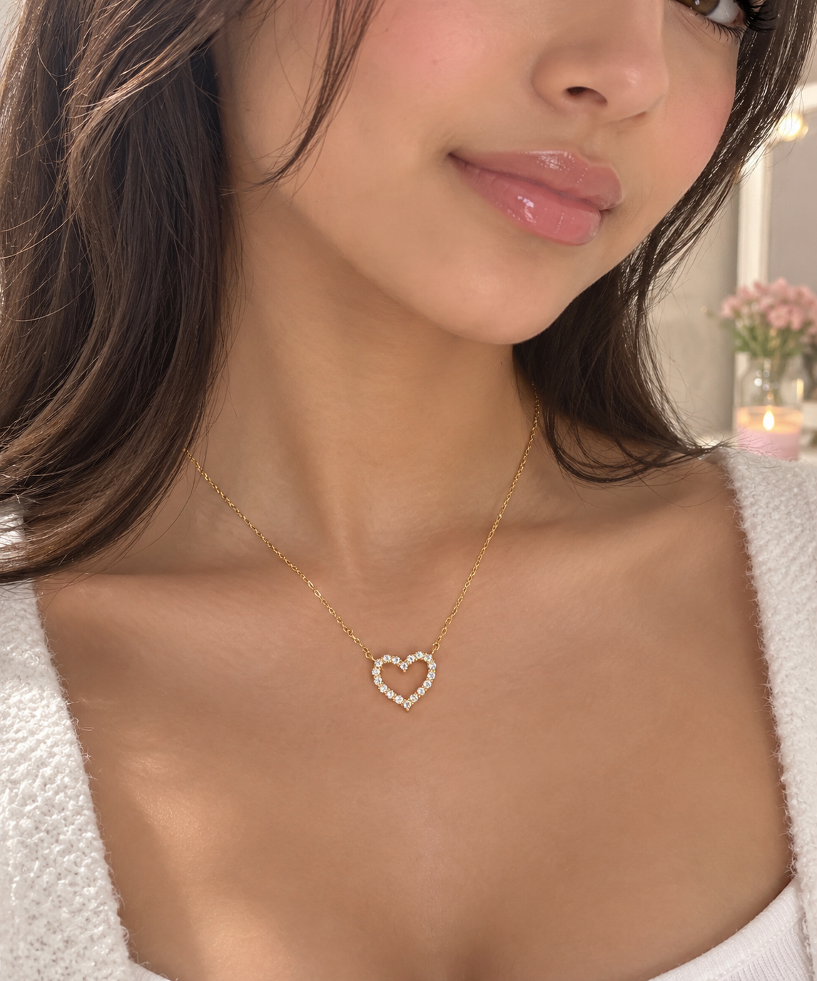 A close-up lifestyle shot of a woman wearing a delicate gold chain necklace with a small, open heart-shaped pendant adorned with tiny clear stones.