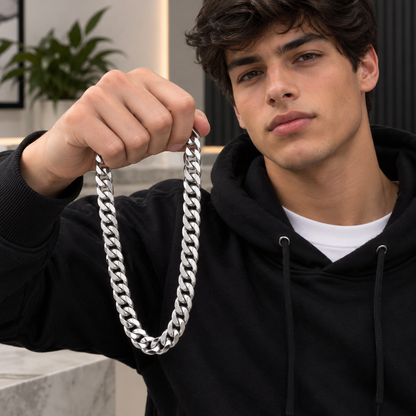 A man wearing a black hoodie holds up a thick, shiny silver Cuban link chain necklace in front of his face. The background shows a modern, softly blurred indoor setting with a plant and light-colored walls.