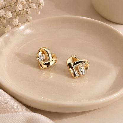  pair of gold heart-shaped stud earrings with small clear crystal clusters, displayed on a round beige ceramic dish with dried baby's breath flowers in the background.