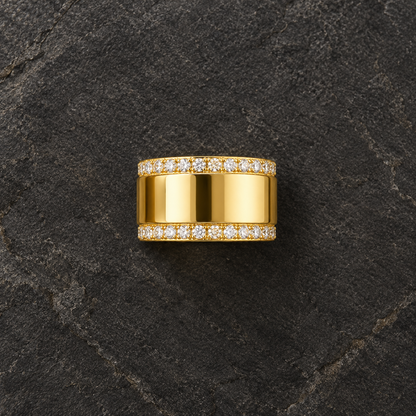 A wide yellow gold ring with pavé-set clear stones on both sides, centered on a dark textured stone surface.