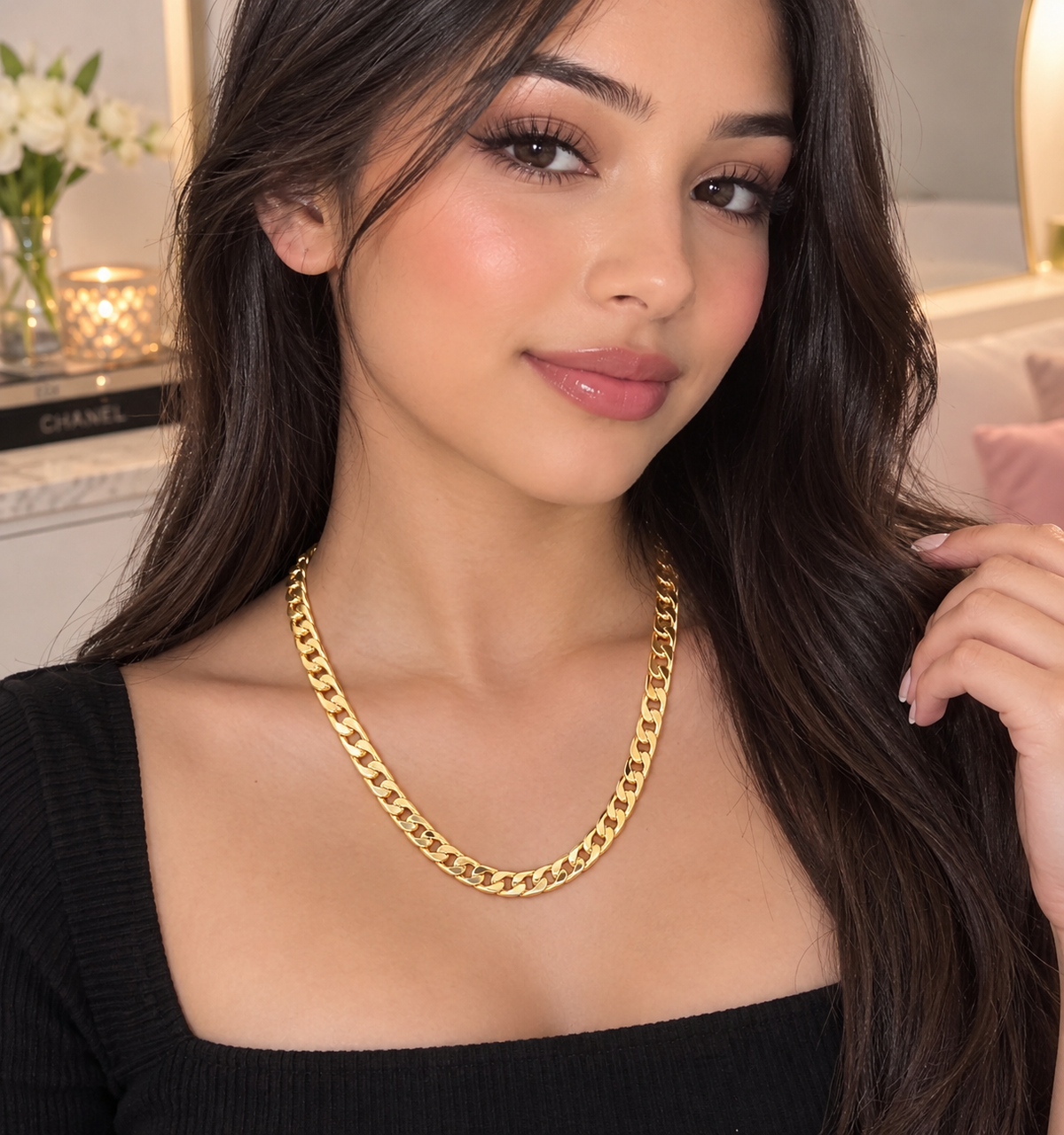 A woman with long dark hair models a thick gold Cuban link chain necklace, wearing a black top in a softly lit indoor setting.