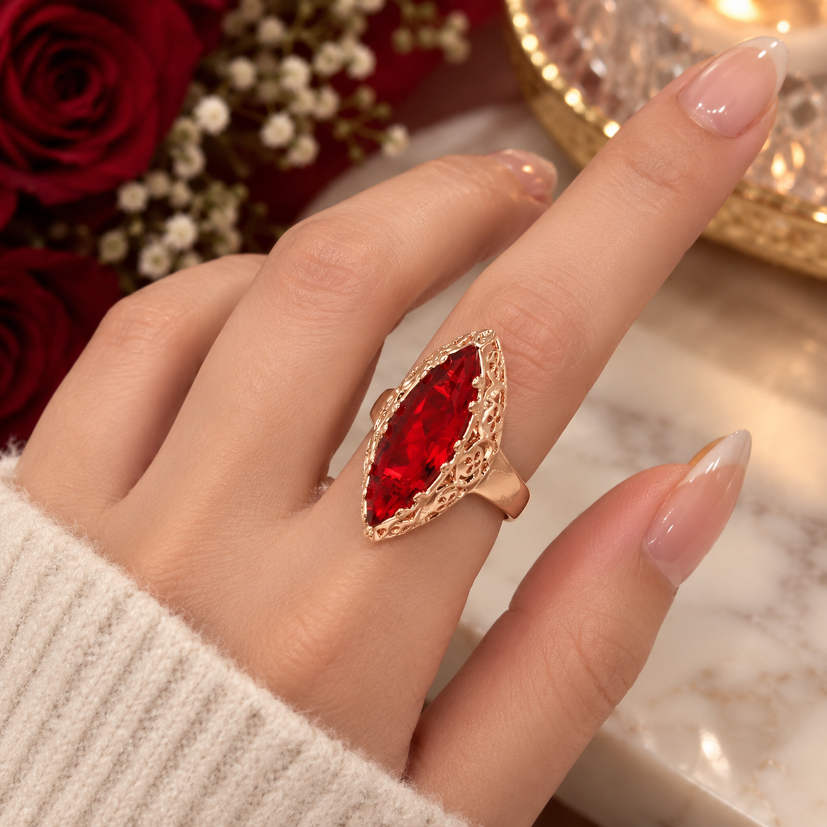 A close-up of a hand wearing a gold ring with a large marquise-cut red gemstone, set against a background of red roses and a marble surface.