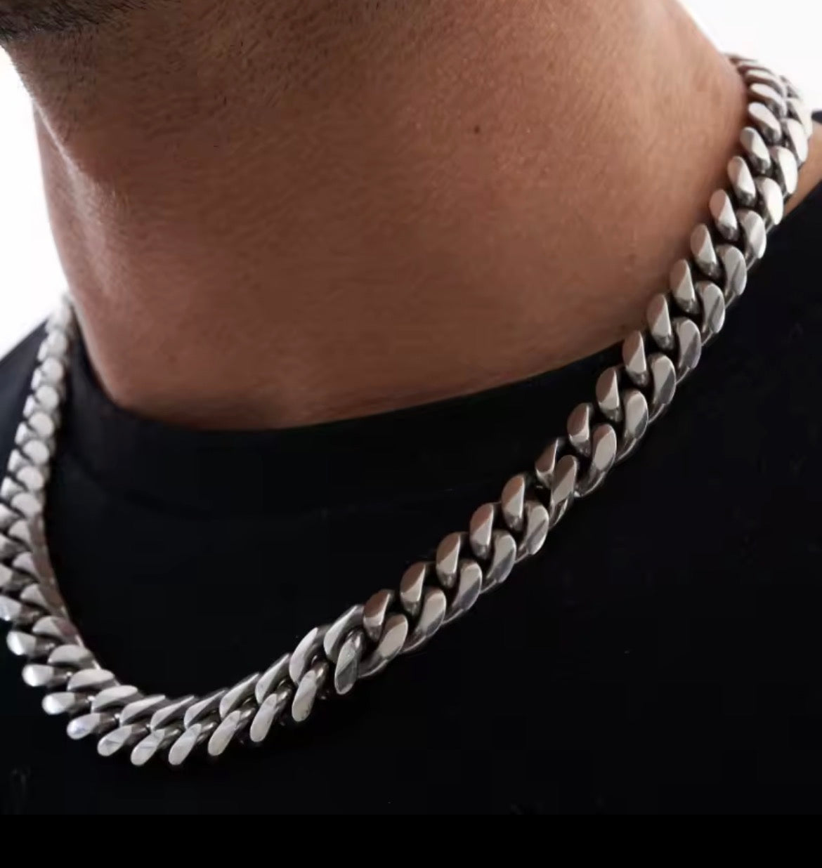A close-up of a man's neck and chest wearing a thick, polished silver Cuban link chain necklace over a black t-shirt.