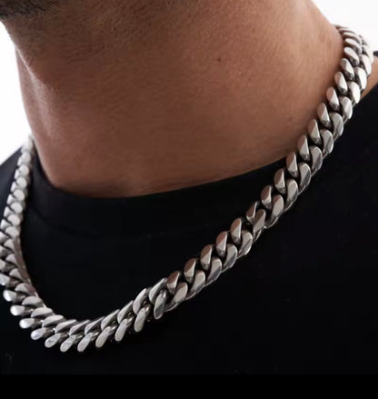 A close-up of a man's neck and chest wearing a thick, polished silver Cuban link chain necklace over a black t-shirt.