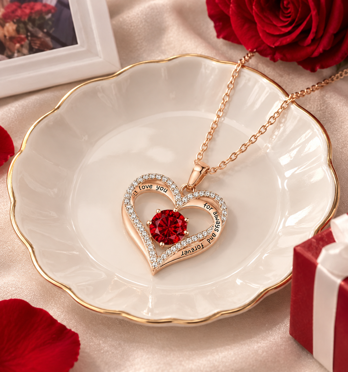 A rose gold heart pendant necklace with a red crystal center and rhinestone-accented border, displayed on a white scalloped dish with gold trim. The necklace is styled with red roses, a gift box, and a framed photo on a satin background.