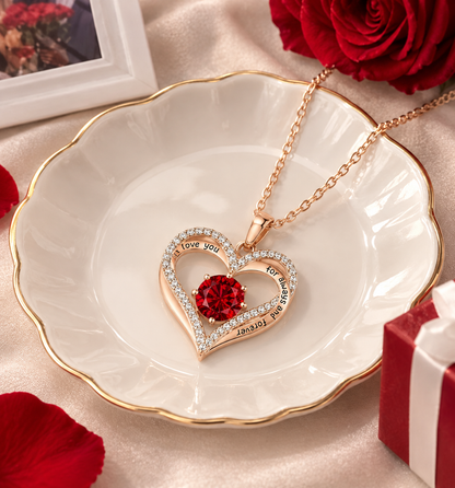 A rose gold heart pendant necklace with a red crystal center and rhinestone-accented border, displayed on a white scalloped dish with gold trim. The necklace is styled with red roses, a gift box, and a framed photo on a satin background.