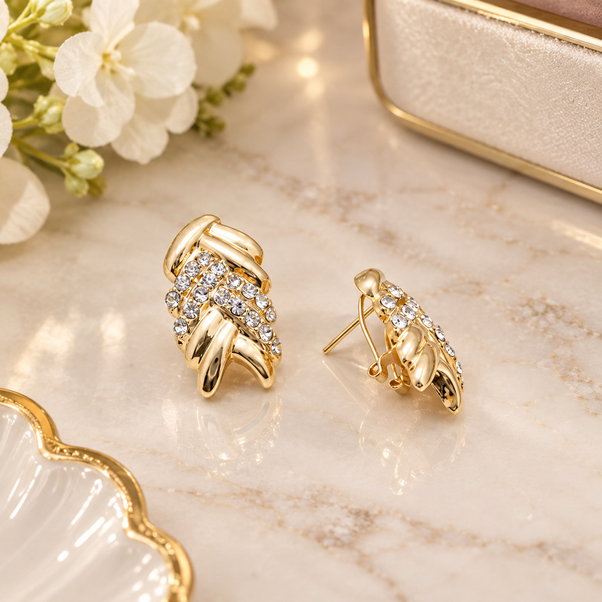 A pair of gold-toned statement earrings with clear crystal clusters, displayed on a light marble surface with white flowers and a cream jewelry box in the background.