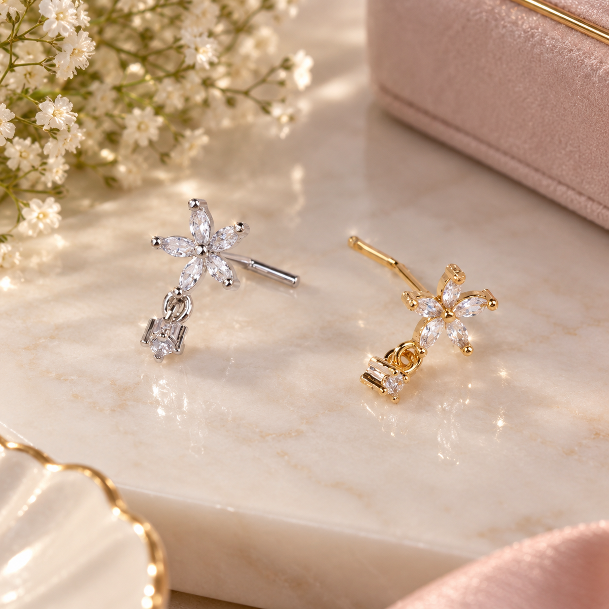 Silver-tone and gold-tone L-shaped nose studs with five-petal clear-stone flowers and dangling clear stones, displayed on a cream surface.
