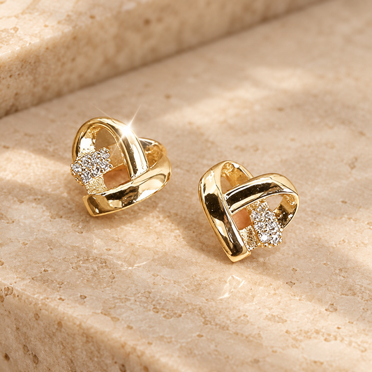 A pair of gold heart-shaped stud earrings with clear crystal accents, displayed on a beige stone surface.