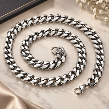 A chunky silver Cuban link chain necklace arranged in a spiral on a light marble surface with a black box corner and small white flowers as styled props
