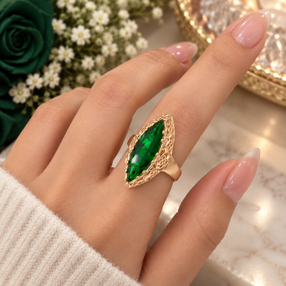 A close-up of a hand wearing a gold ring with a marquise-cut green emerald gemstone, set against a soft-focus background of flowers and a marble surface.