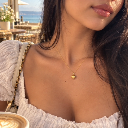A close-up lifestyle shot of a woman wearing a delicate gold heart pendant necklace, styled with a white ruffled top and a gold chain bag strap, set against a warm, sunlit outdoor cafe background with a hint of the ocean.

