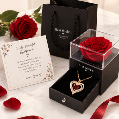 A romantic gift set featuring a red rose preserved in a clear display box with a heart-shaped gold pendant, a black gift bag, and a printed message card on a white marble surface.