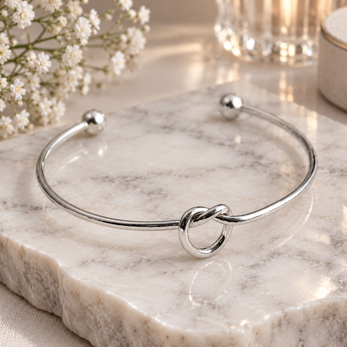 A polished silver open cuff bracelet with a central knotted ring detail, displayed on a white marble slab with small white flowers in the background.