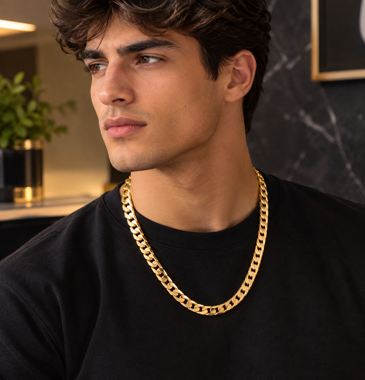 A man wearing a thick, shiny gold Cuban link chain necklace over a black t-shirt, photographed in a modern indoor setting with a dark marble wall background.