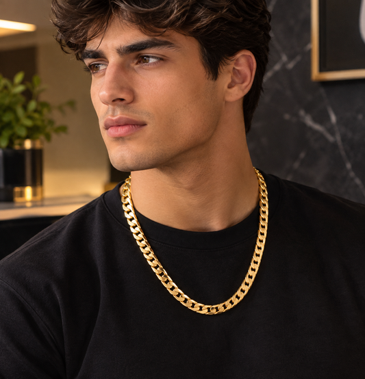 A man wearing a thick, shiny gold Cuban link chain necklace over a black t-shirt, photographed in a modern indoor setting with a dark marble wall background.