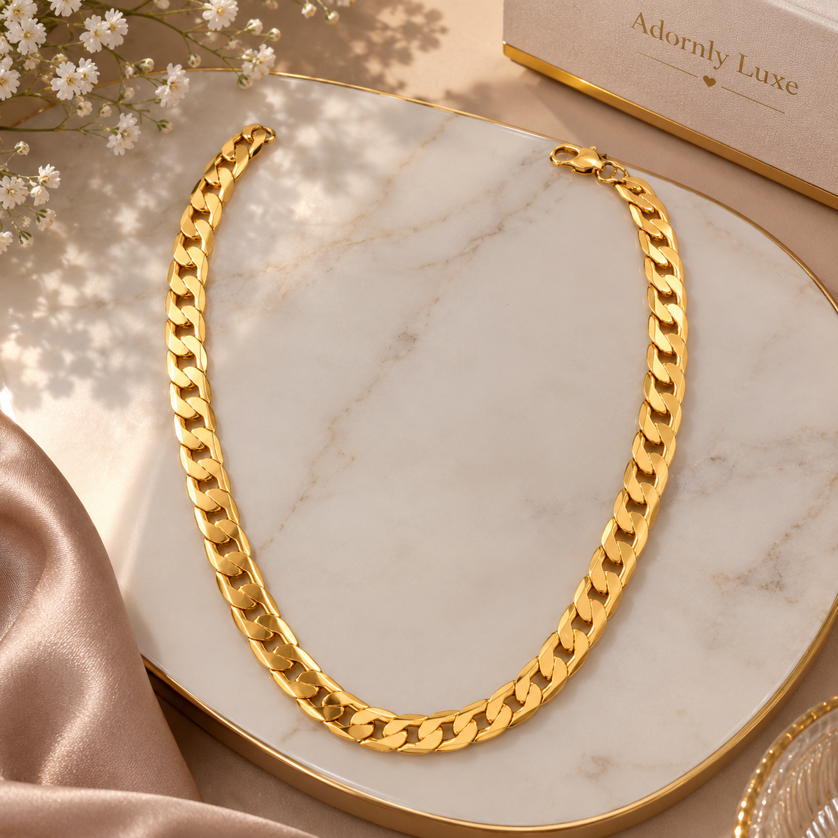 A thick gold Cuban link chain necklace displayed on a white marble tray with gold trim, styled with satin fabric, small white flowers, and a cream jewelry box labeled Adornly Luxe.