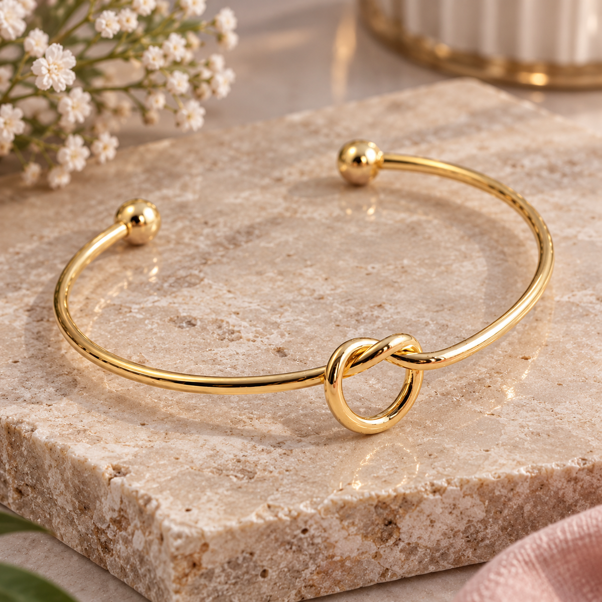 A polished gold open cuff bracelet with a central knotted ring detail, displayed on a textured beige stone slab with small white flowers in the background.