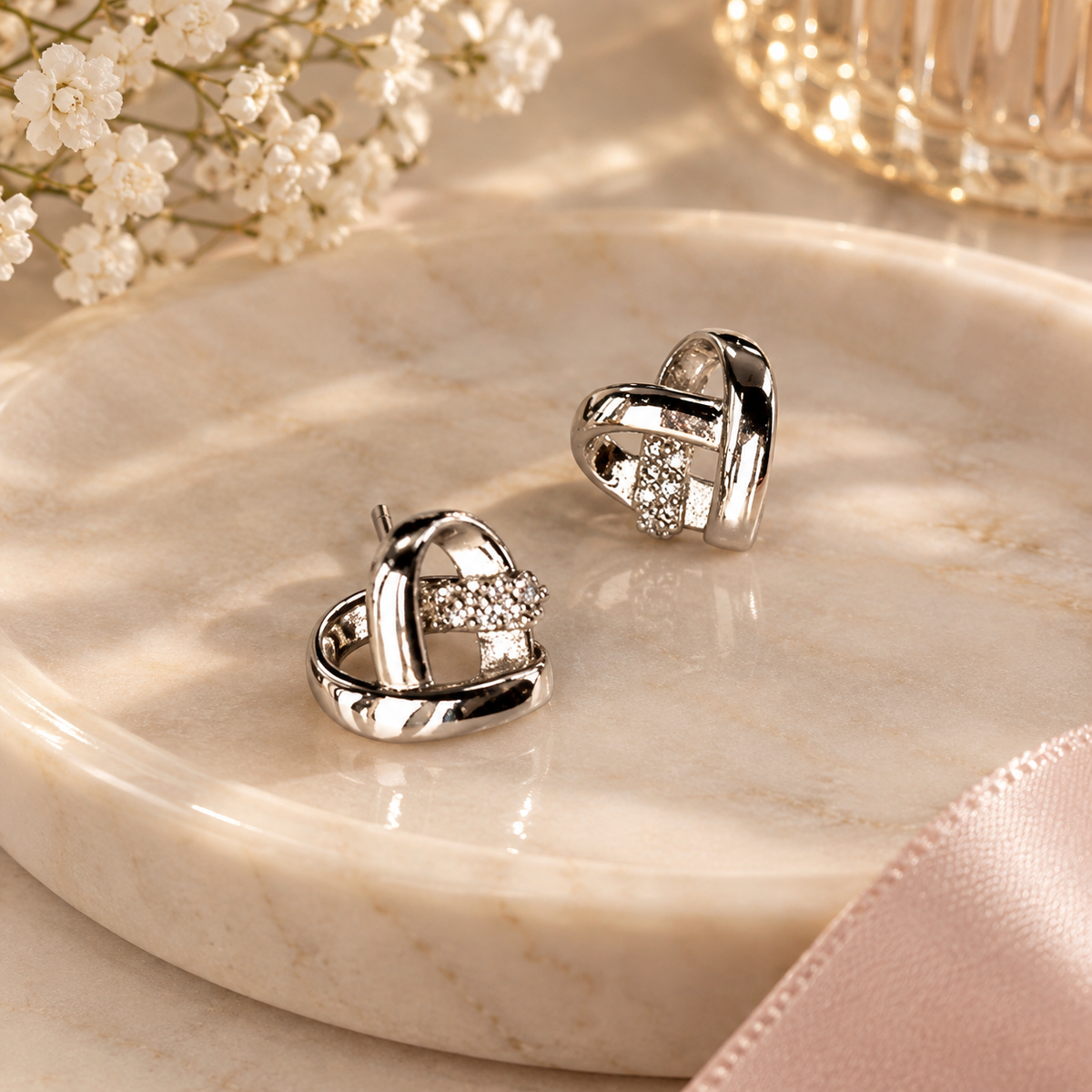 A pair of polished silver heart-shaped earrings with small clear crystal accents, displayed on a round beige marble tray with soft floral and ribbon props.