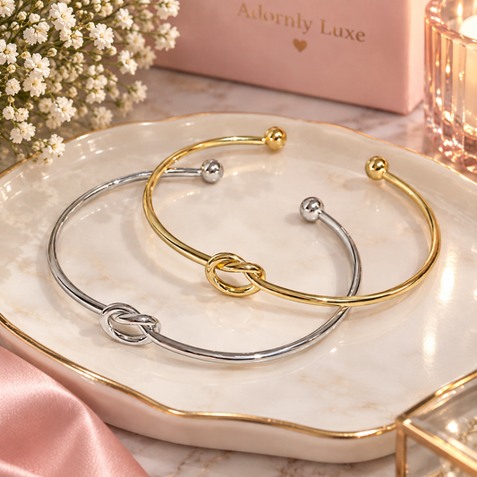 Two open cuff bracelets, one in polished gold and one in polished silver, both featuring a central knot design and small bead accents, displayed on a white marble tray with gold trim.
