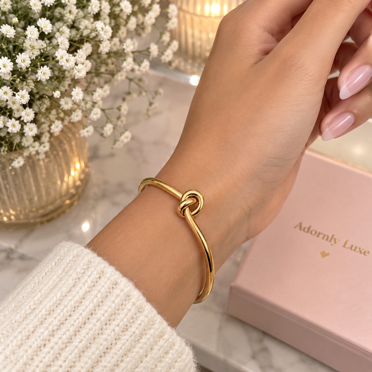 A gold-toned bangle bracelet with a knotted center detail worn on a person's wrist, shown in a styled indoor setting with a pink jewelry box labeled 'Adornly Luxe' and white flowers in the background.