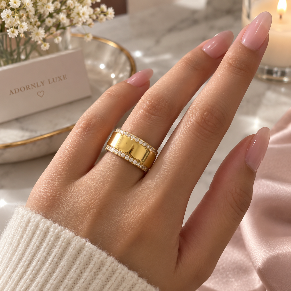 A wide gold ring with two rows of small clear stones, show it on a model engagement finger The scene includes soft pink fabric, white baby's breath flowers, and a brand card in display. 